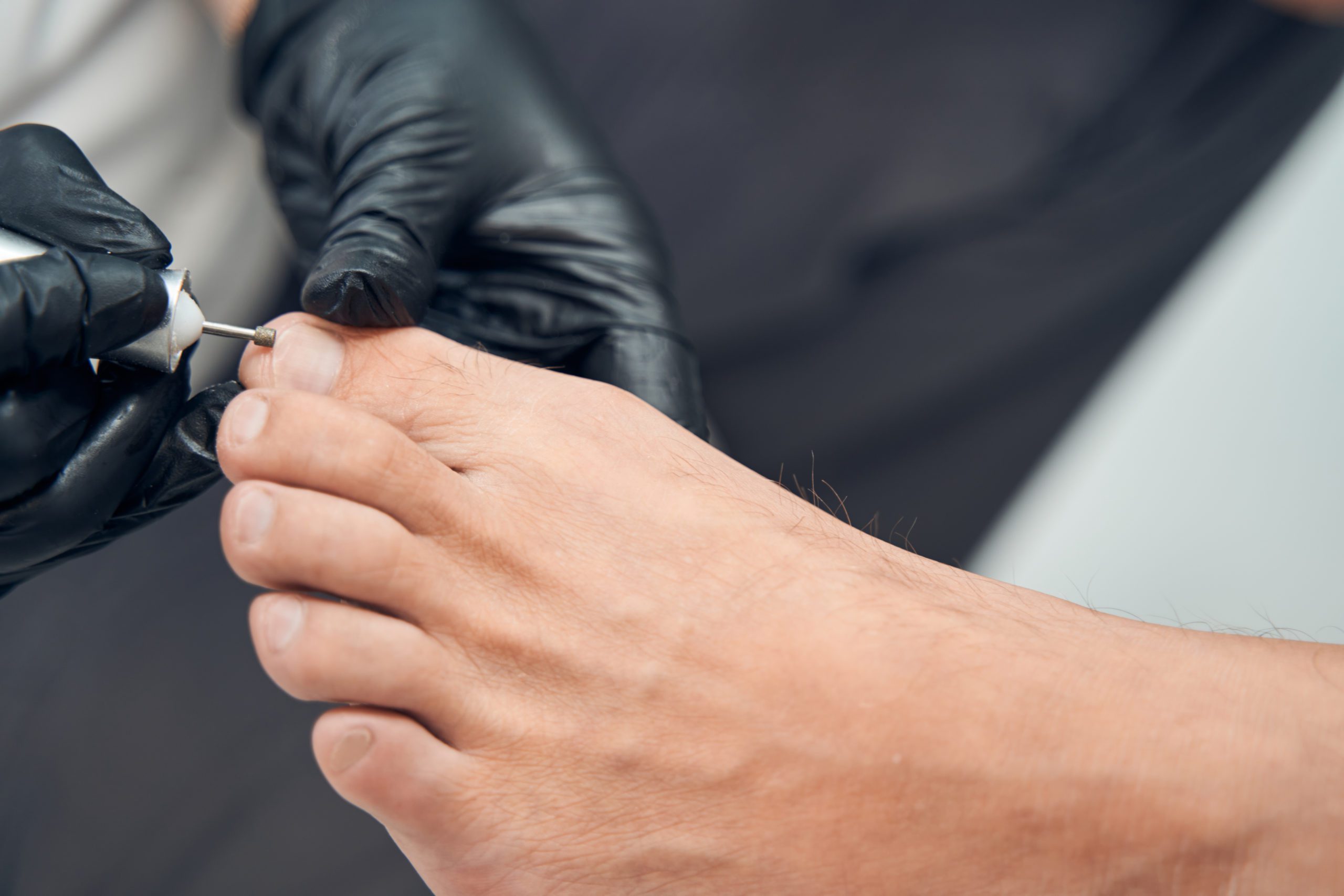 Beautician hands doing hardware pedicure for male client Close up of female specialist in sterile gloves using electric device while doing pedicure for man in cosmetology salon